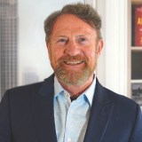 A middle-aged man with a beard and mustache, wearing a blue suit jacket and light blue shirt, standing indoors with bookshelves and a window in the background.