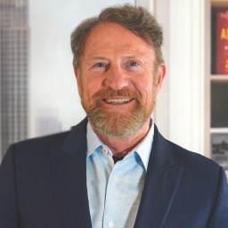 A middle-aged man with a beard and mustache, wearing a blue suit jacket and light blue shirt, standing indoors with bookshelves and a window in the background.