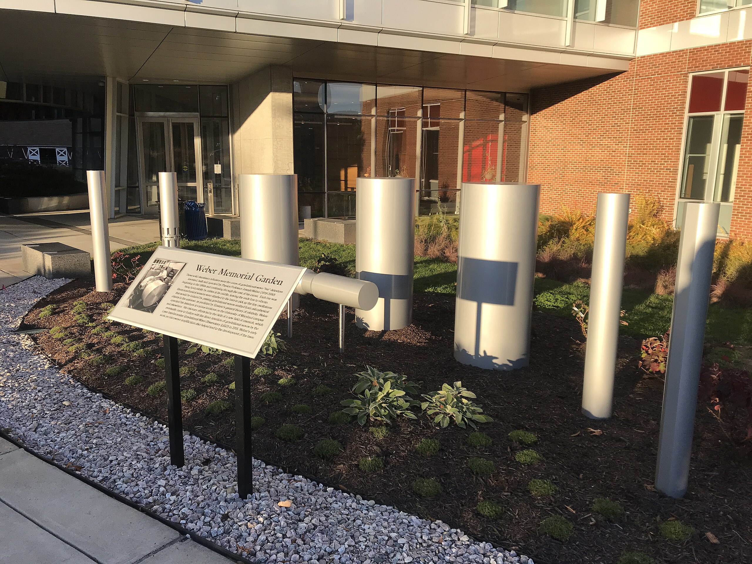 An outdoor garden features metal cylindrical sculptures reminiscent of Weber bars used in detecting gravitational waves, a sign with information, and landscaping with rocks and small plants in front of a modern building.