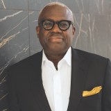 A man wearing glasses, a dark suit with a yellow pocket square, and a white shirt stands in front of a marble wall.