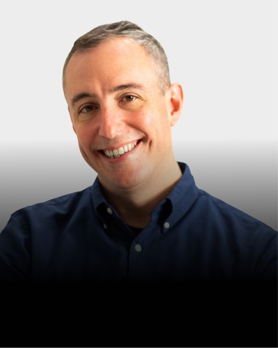 A man with short gray hair, wearing a navy blue button-up shirt, smiles at the camera against a light gray background.