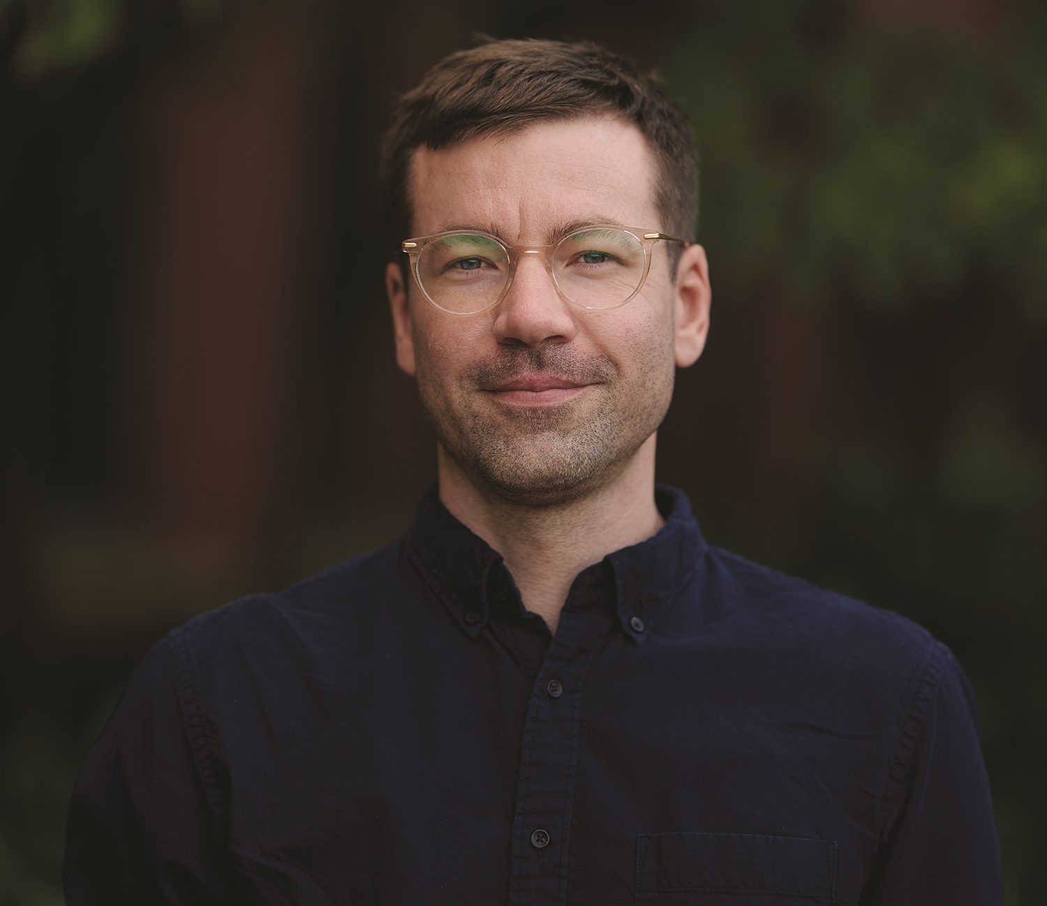 A man with short brown hair and glasses, wearing a dark button-up shirt, stands outdoors in front of a blurred green background.