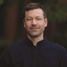 A man with short brown hair and glasses, wearing a dark button-up shirt, stands outdoors in front of a blurred green background.