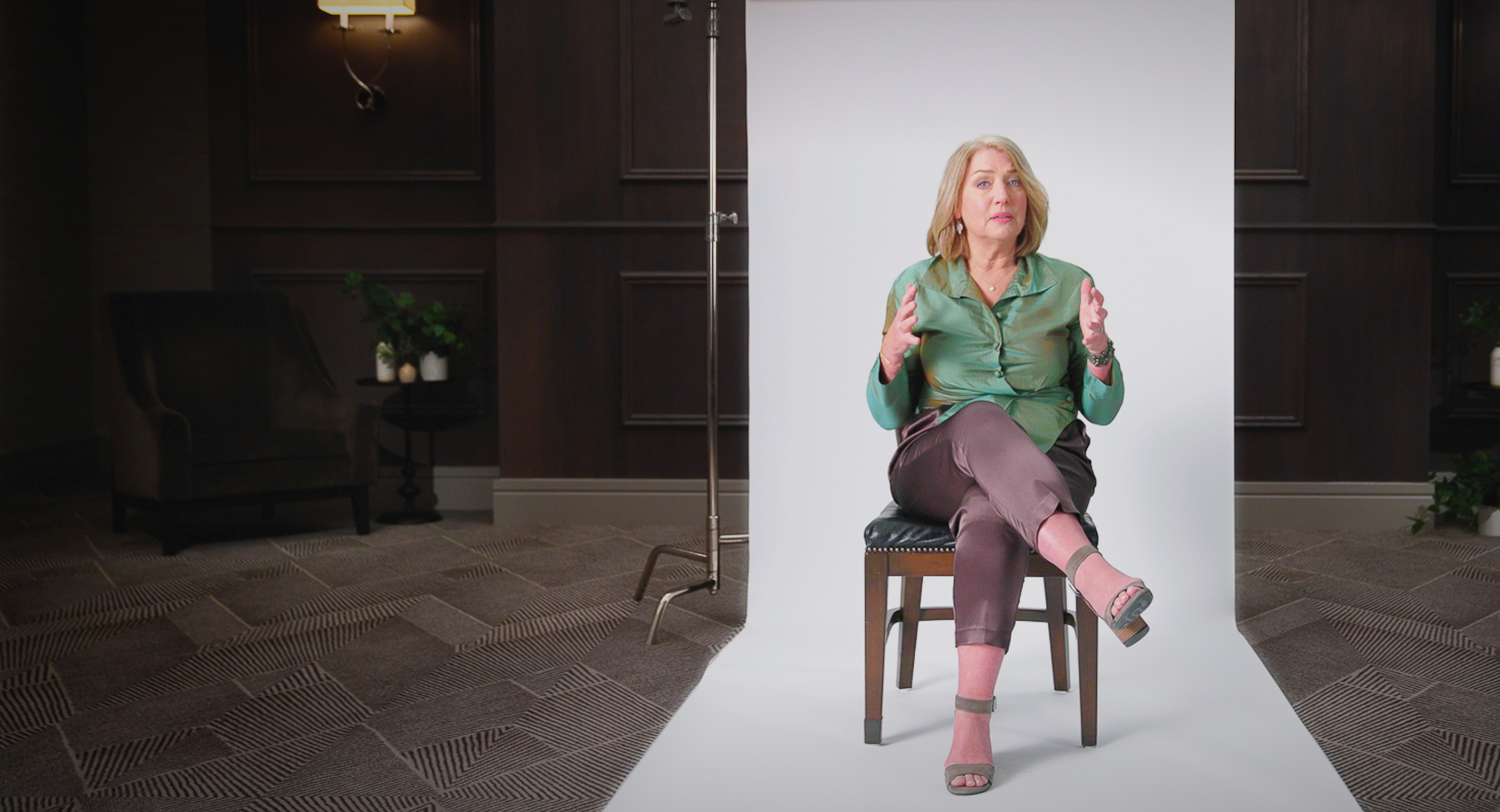 A woman in a green blouse and brown pants sits on a chair, gesturing with her hands, in front of a white backdrop in a dimly lit room.