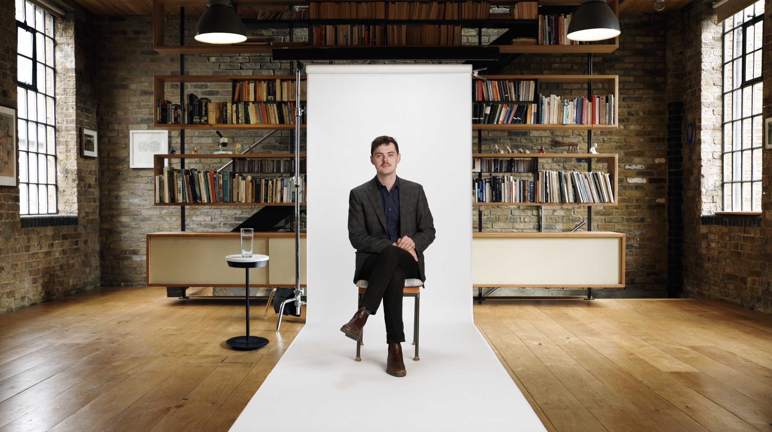 A man sits on a chair in front of a white backdrop in a library with brick walls, wooden floor, shelves of books, and large windows.