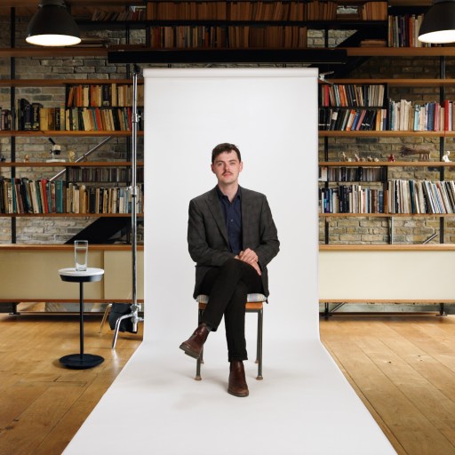 A man sits on a chair in front of a white backdrop in a library with brick walls, wooden floor, shelves of books, and large windows.