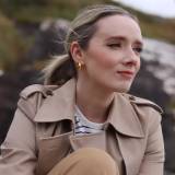 A woman with blonde hair tied back, wearing a beige trench coat and gold earrings, sits outdoors with a neutral expression.