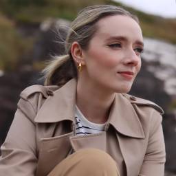 A woman with blonde hair tied back, wearing a beige trench coat and gold earrings, sits outdoors with a neutral expression.