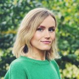 A person with shoulder-length blonde hair wearing a green sweater stands outdoors in front of a blurred green leafy background.