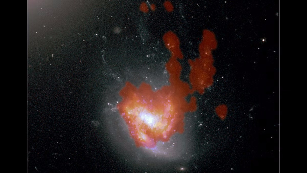 A jellyfish galaxy with a bright white and yellow core is surrounded by red clouds of gas, set against a dark background filled with stars.