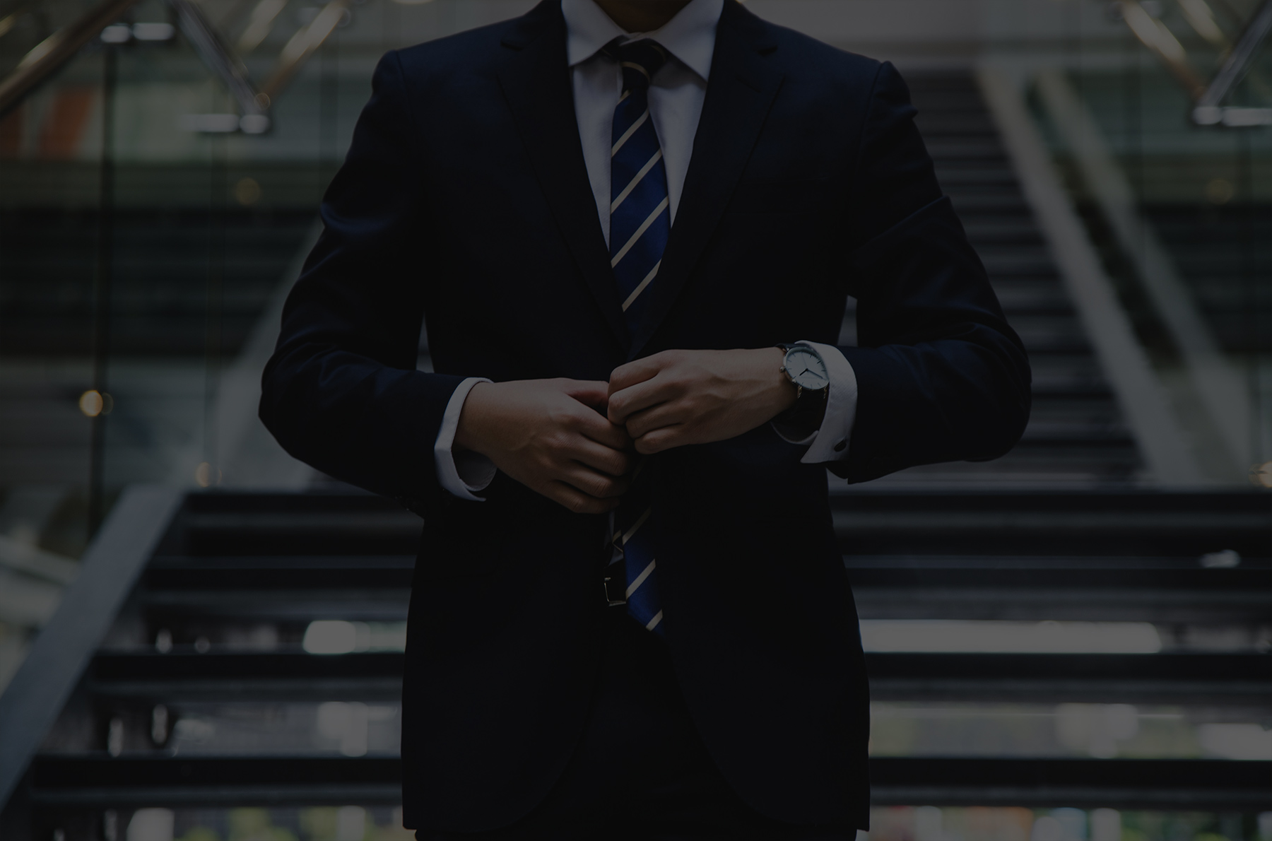 A person in a dark suit and striped tie buttons their jacket, standing in front of a staircase in a modern building.