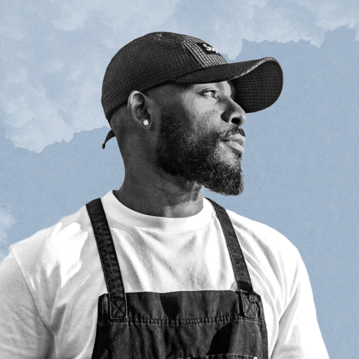 Davon Moseley, a man with a beard wearing a white shirt, black apron, and a cap, stands against a blue sky with clouds.