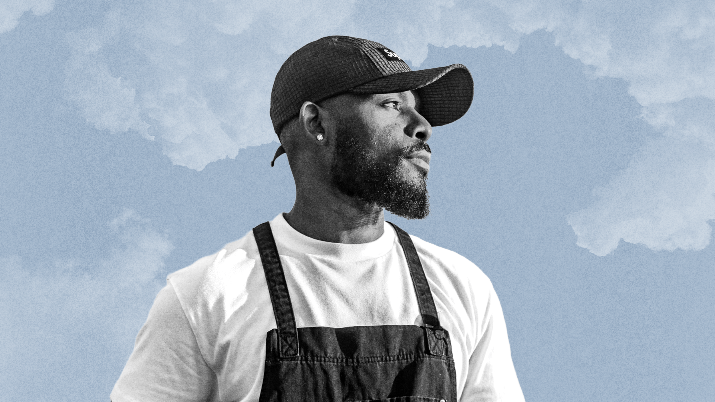 Davon Moseley, a man with a beard wearing a white shirt, black apron, and a cap, stands against a blue sky with clouds.