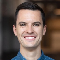 A man with short dark hair and a blue patterned shirt smiles at the camera against a blurred indoor background.