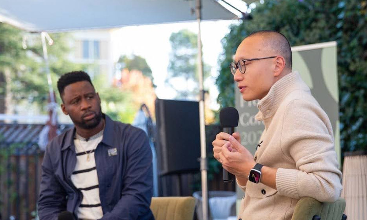 Two men sit outdoors; one speaks into a microphone while the other listens attentively. There is greenery and a blurred banner in the background.