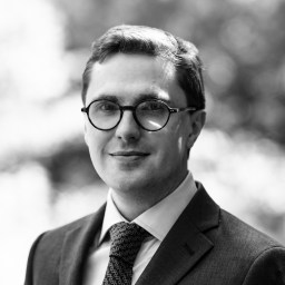 A man wearing glasses, a suit, and a patterned tie looks at the camera with a neutral expression. The background is blurred.