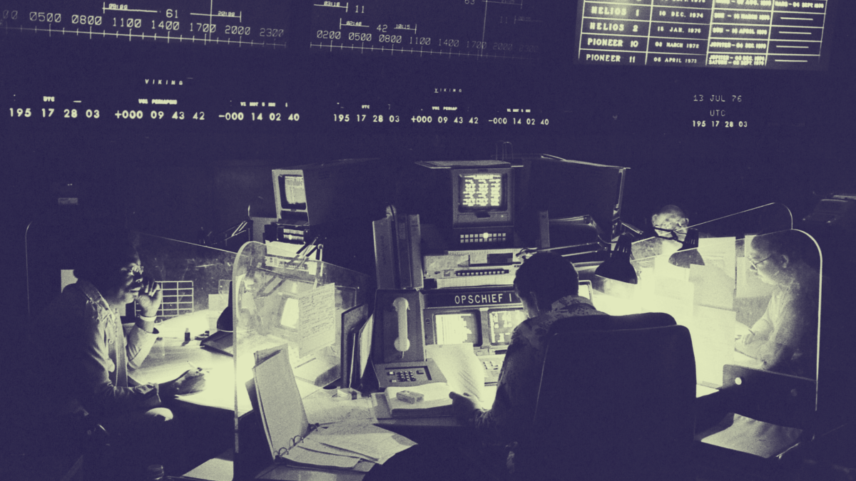 Four people work at consoles surrounded by monitors and control panels in a dimly lit NASA mission control room, with large display boards overhead.