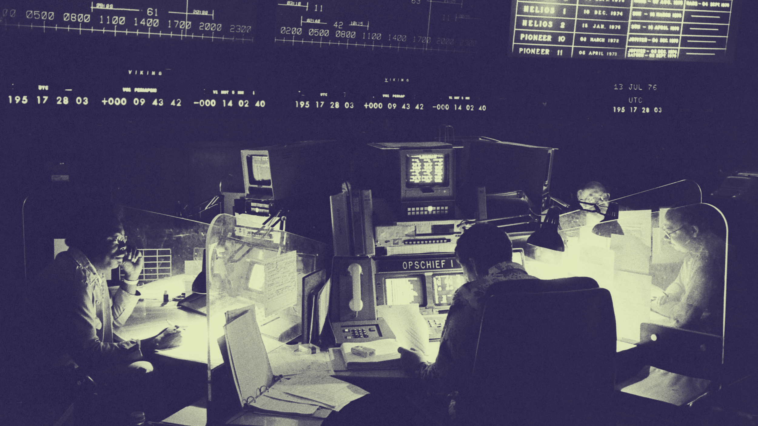 Four people work at consoles surrounded by monitors and control panels in a dimly lit NASA mission control room, with large display boards overhead.