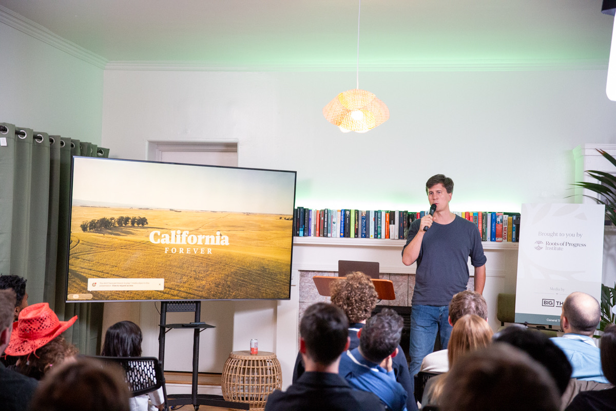 A man stands with a microphone addressing an audience in a room; a large screen displays "California Forever" next to him.