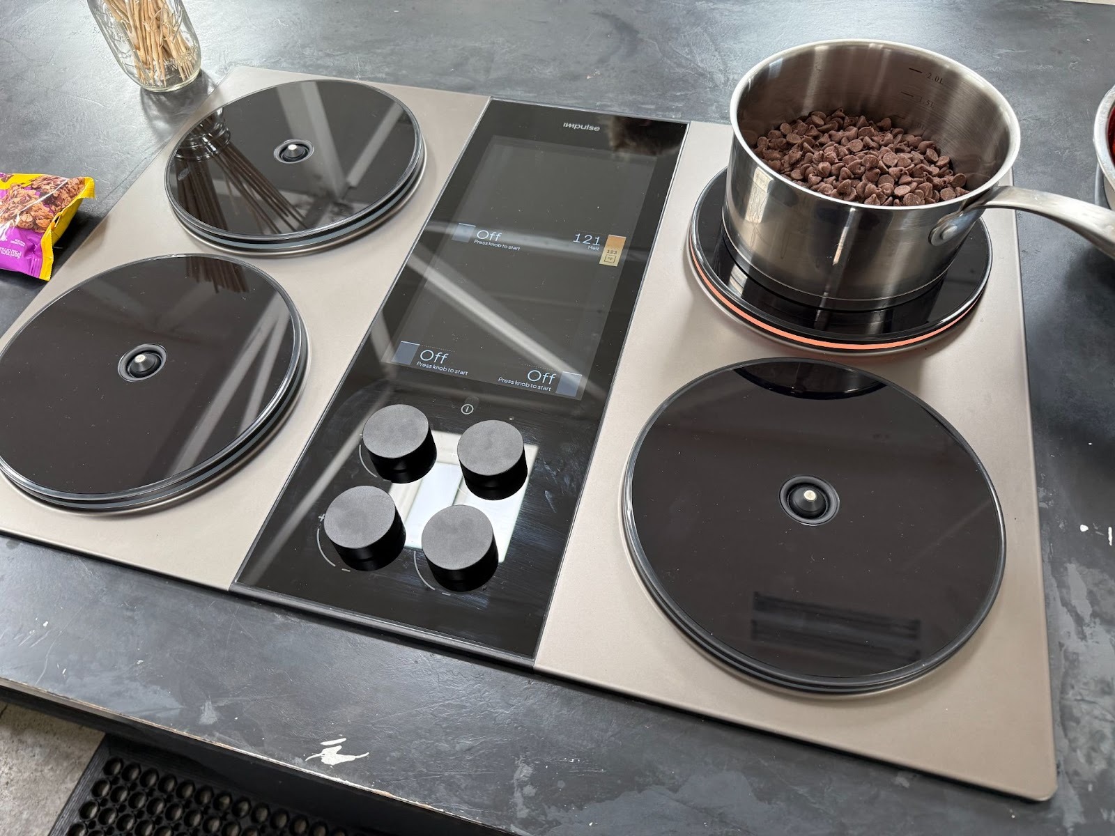 A pot filled with chocolate chips is melting on an electric stove with four burners; three burners are covered with lids.
