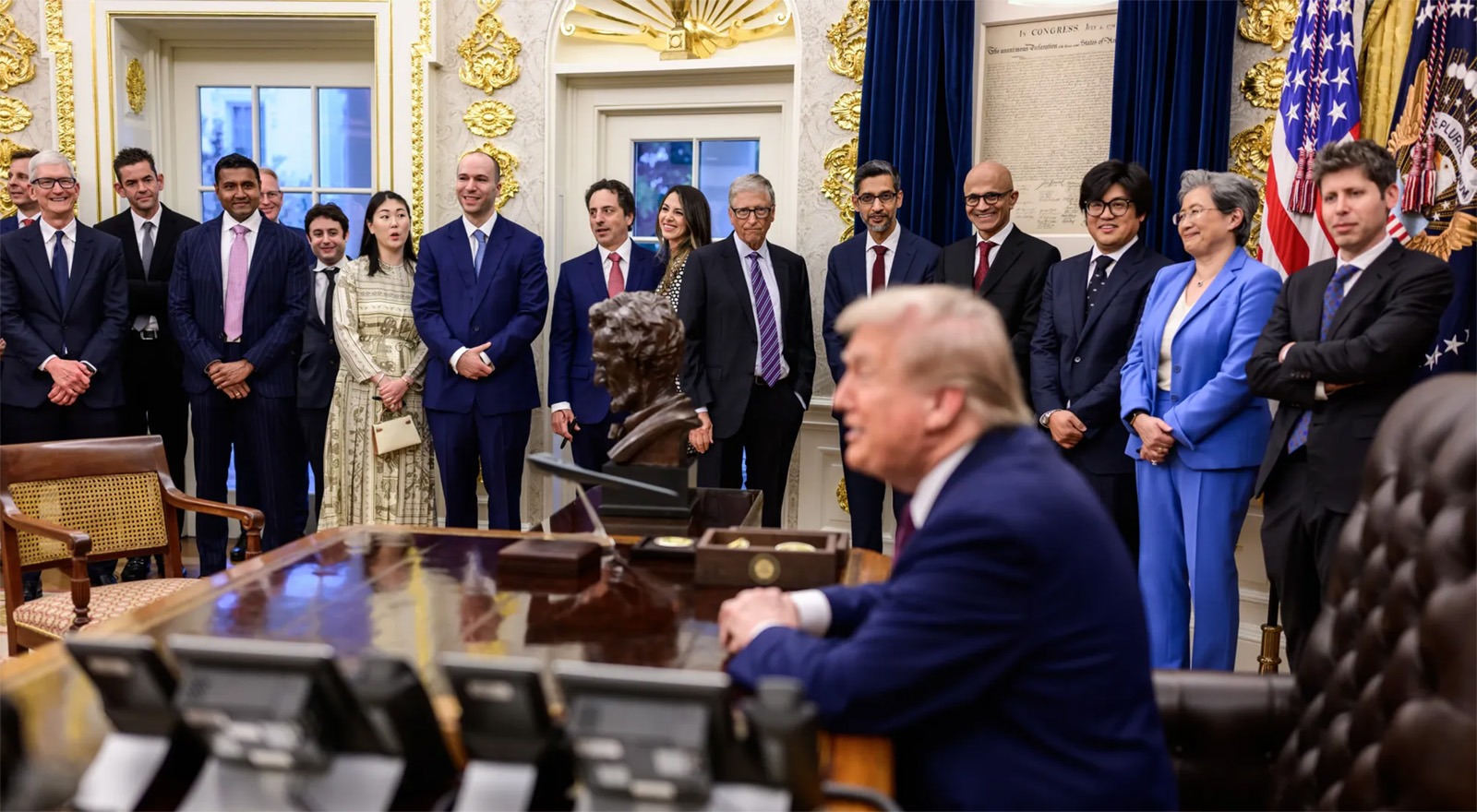 A group of people in business attire stand facing a man seated at a desk in an ornate office with gold decor and dark blue curtains.