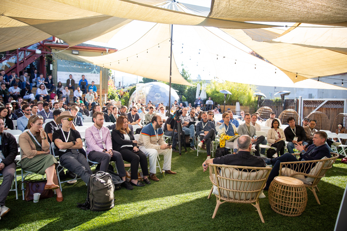 A large group of people seated outdoors under sunshades, attentively listening to a panel discussion with two speakers seated in wicker chairs at the front.