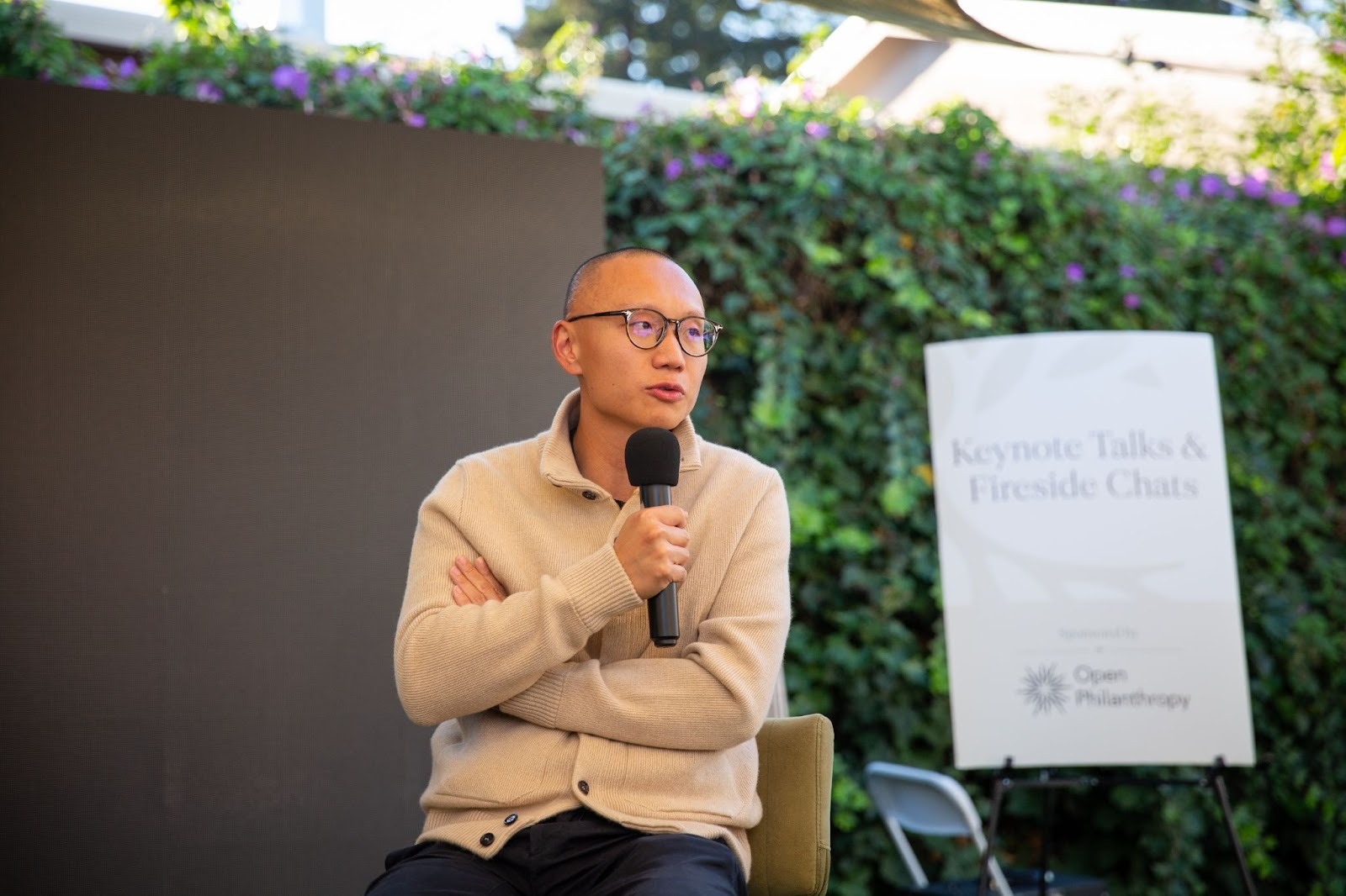 A person in glasses and a beige sweater sits holding a microphone, speaking at an outdoor event. A sign in the background reads "Keynote Talks & Fireside Chats, Open Philanthropy.