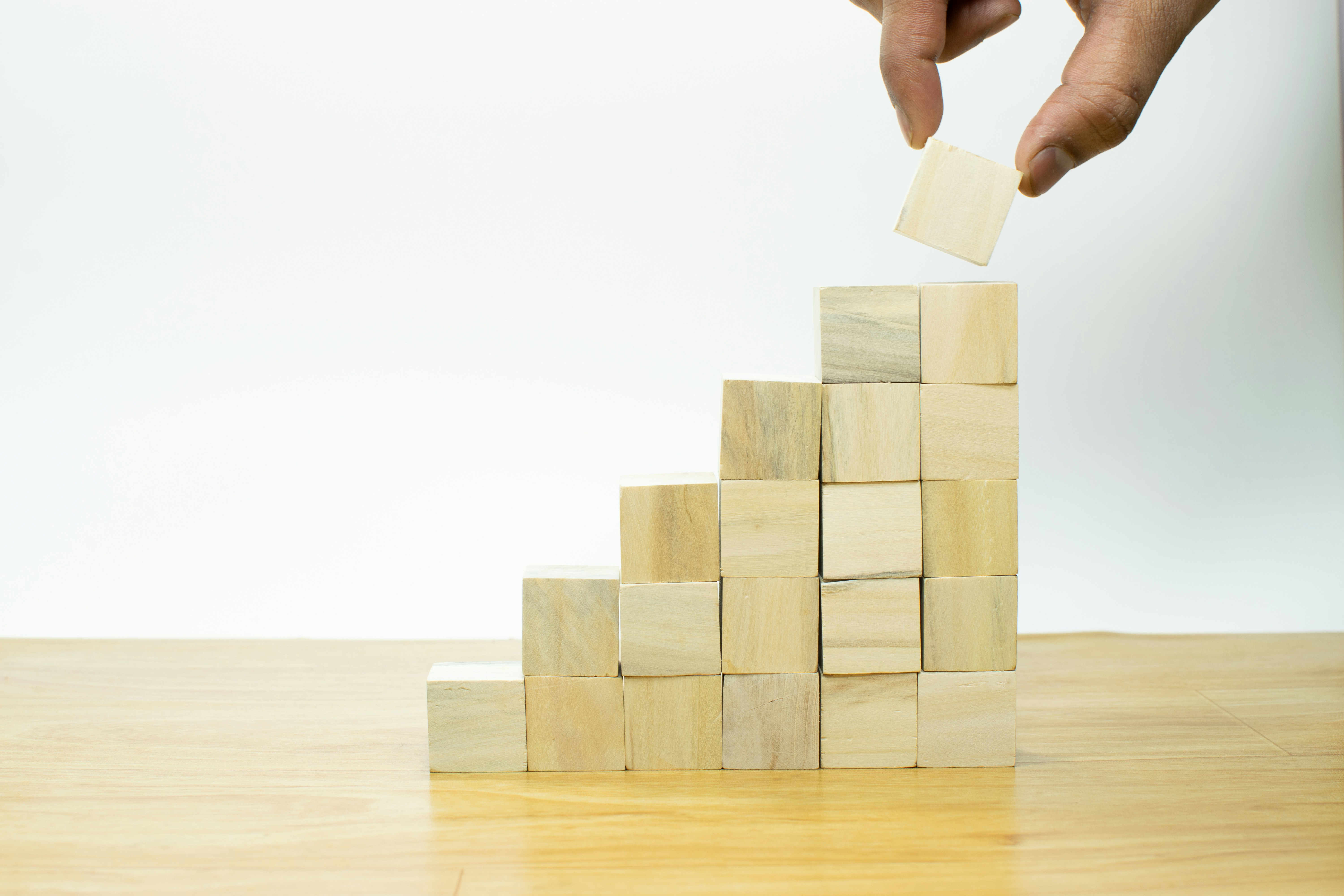 A hand places a wooden block on top of a staircase-like structure made of wooden blocks on a wooden surface.