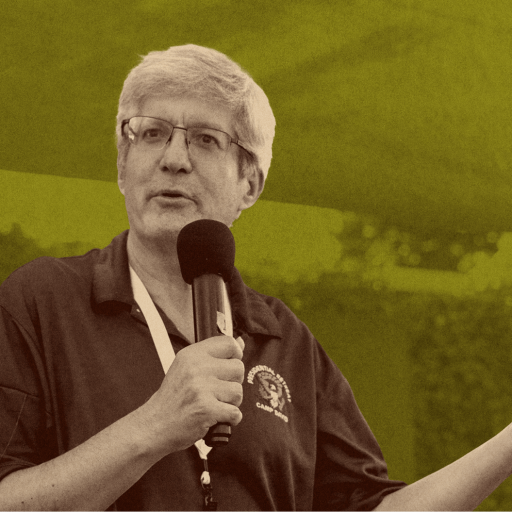 An older man with gray hair and glasses speaks into a microphone, gesturing with one hand, against a green and grid-patterned background.