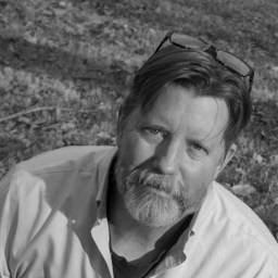 A middle-aged man with a beard and mustache, wearing glasses on his head and a button-up shirt, looks up outdoors. The photo is in black and white.