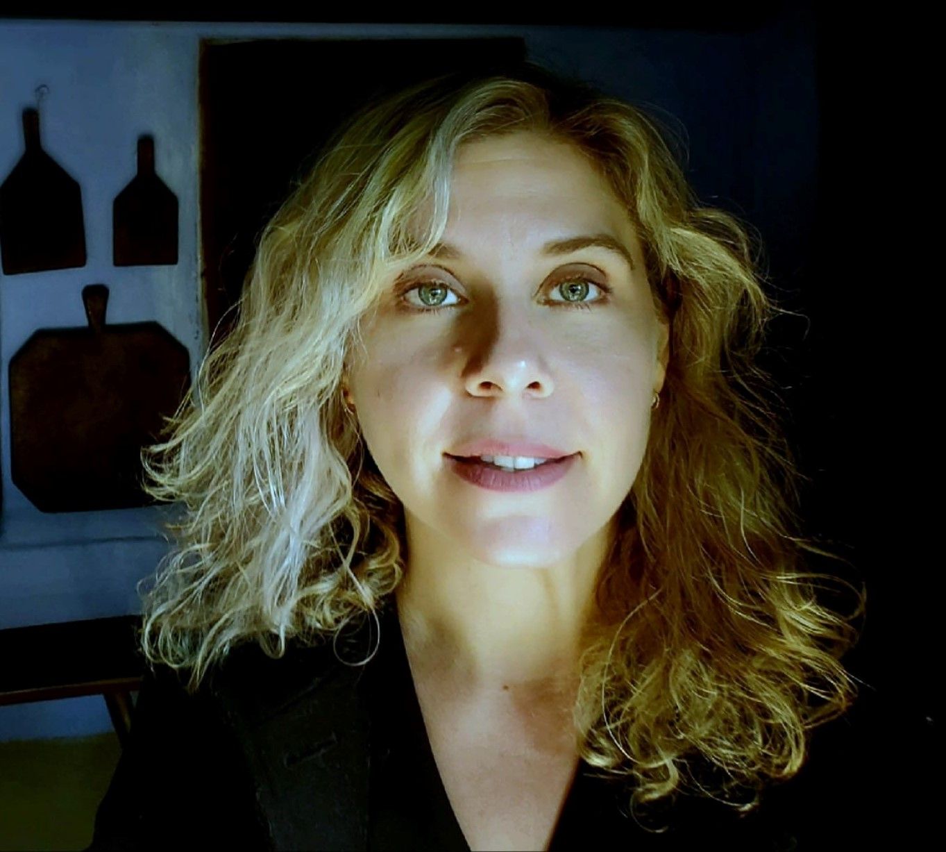 A woman with wavy blonde hair and green eyes is facing the camera in a dimly lit room with cutting boards hanging on the wall in the background.