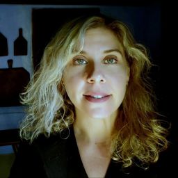 A woman with wavy blonde hair and green eyes is facing the camera in a dimly lit room with cutting boards hanging on the wall in the background.