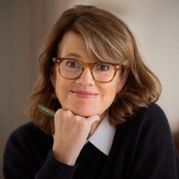 A woman with shoulder-length brown hair and glasses, wearing a black sweater over a white shirt, rests her chin on her hand and holds a pen, looking at the camera.