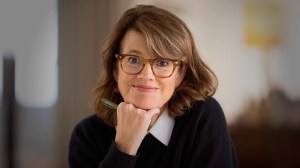 A woman with shoulder-length brown hair and glasses, wearing a black sweater over a white shirt, rests her chin on her hand and holds a pen, looking at the camera.
