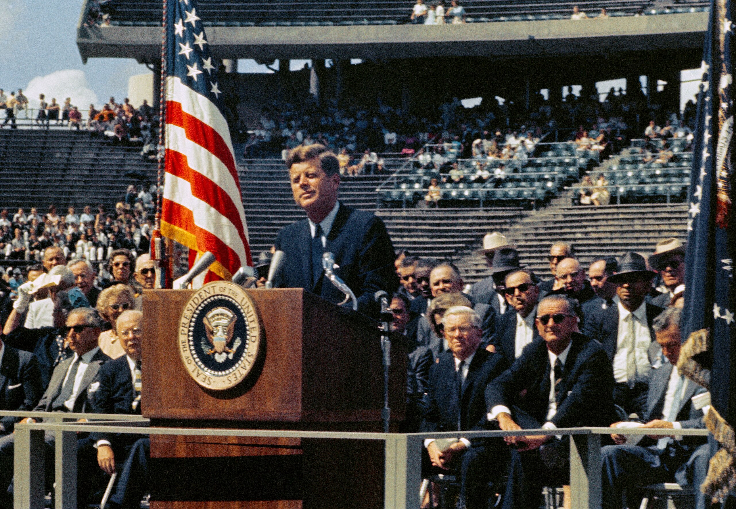 A man in a suit speaks at a podium with the presidential seal, flanked by the U.S. flag, in a stadium with an audience in the background.