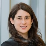 A woman with long brown hair, wearing a black blazer and polka dot blouse, smiles at the camera; background shows blurred vertical panels.