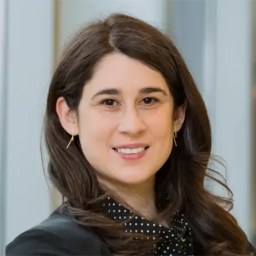 A woman with long brown hair, wearing a black blazer and polka dot blouse, smiles at the camera; background shows blurred vertical panels.
