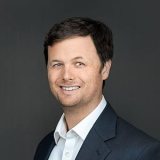 A man with short brown hair wearing a dark suit jacket and a white collared shirt smiles in front of a dark gray background.
