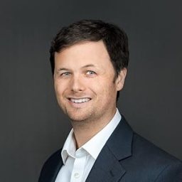 A man with short brown hair wearing a dark suit jacket and a white collared shirt smiles in front of a dark gray background.