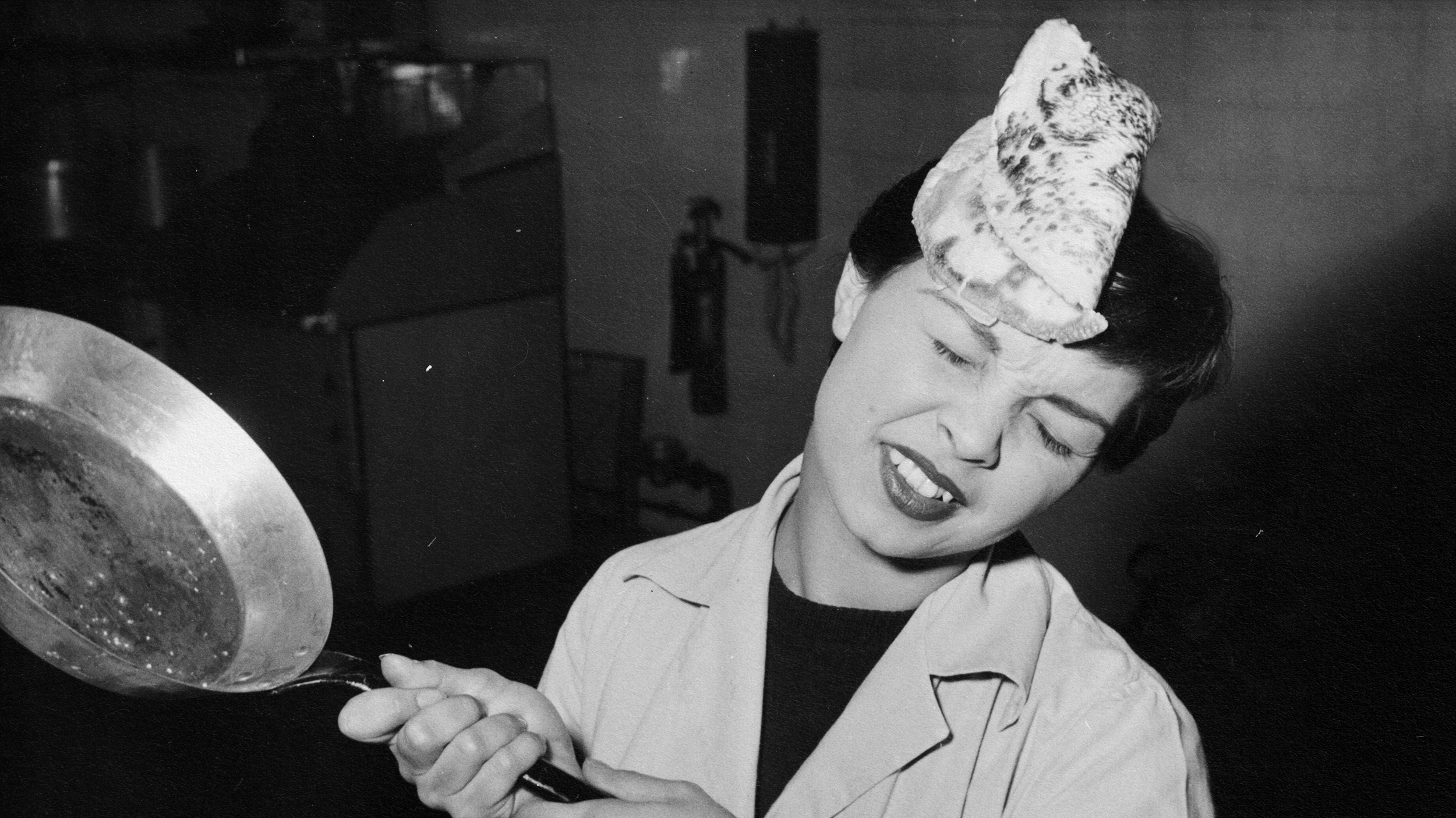 A woman displays stoicism as she grimaces, unfazed by a pancake landing on her head while holding a frying pan in a kitchen setting.