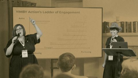 Two women stand and speak in front of a projector screen displaying a graph titled "YIMBY Action’s Ladder of Engagement" at a presentation or workshop.