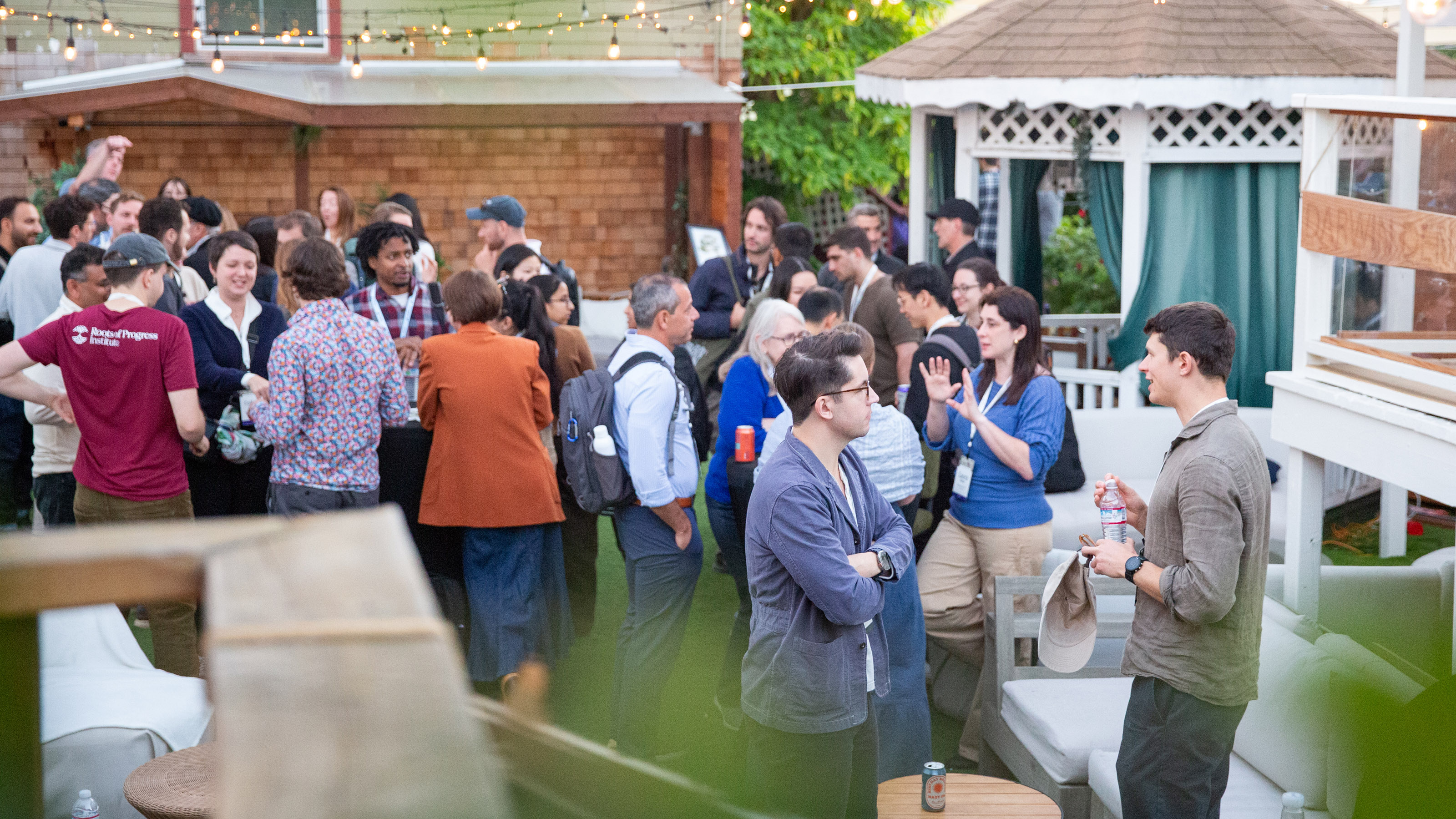 A group of people socialize at an outdoor gathering, standing and conversing in a backyard setting with string lights and a gazebo.