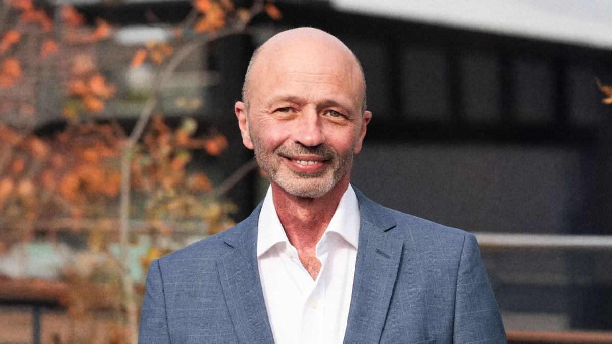 A bald man in a blue suit and white shirt stands outdoors in Silicon Oasis, smiling, with autumn leaves and a blurred building in the background.