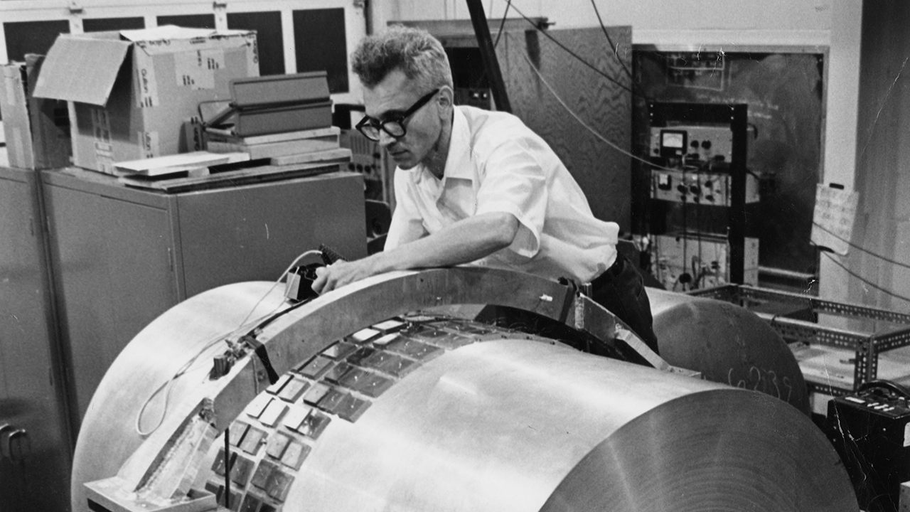 A man adjusts components on a large, cylindrical piece of scientific equipment&mdash;resembling early Weber bars used to detect gravitational waves&mdash;in a laboratory setting.