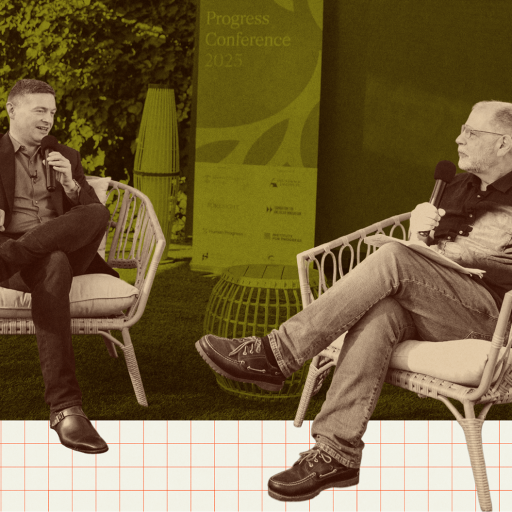Two men sit on outdoor chairs holding microphones, engaged in conversation at an event. A conference sign is visible in the background.