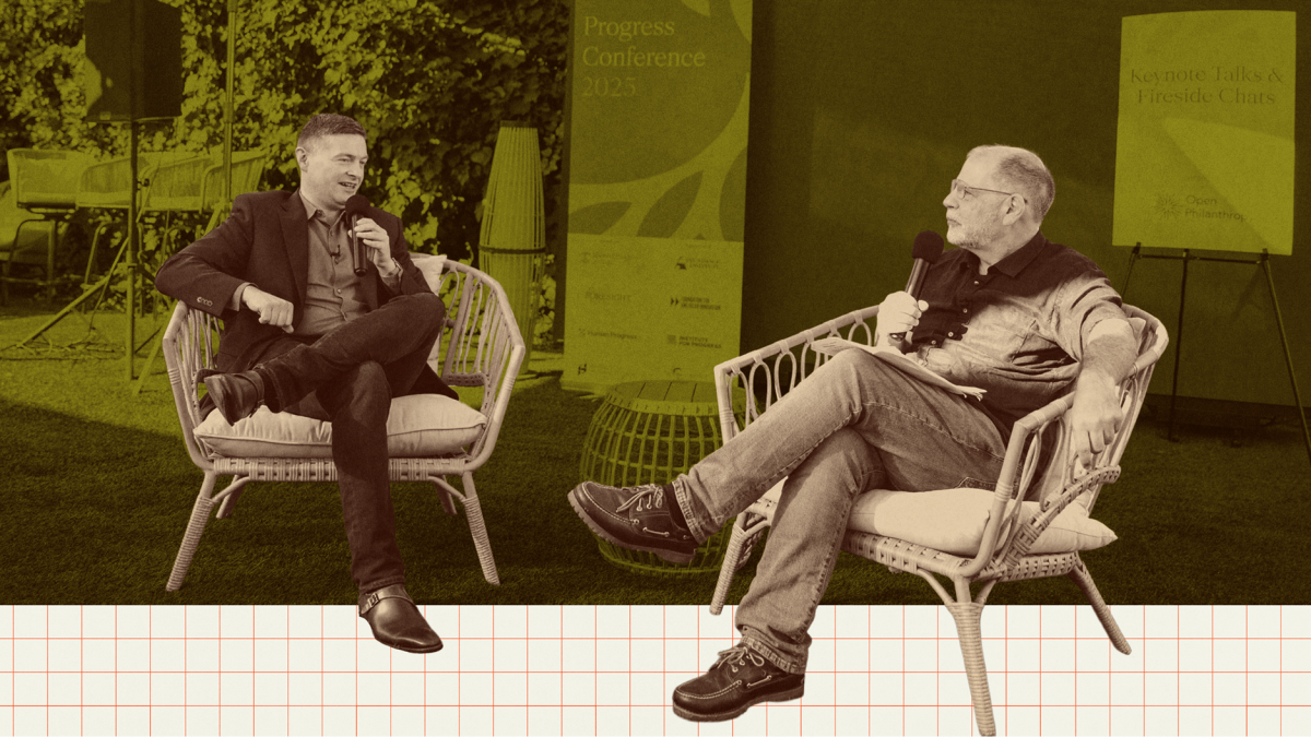 Two men sit on outdoor chairs holding microphones, engaged in conversation at an event. A conference sign is visible in the background.