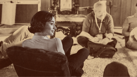 A woman sits in a chair facing a group of people seated on the floor in a casual indoor setting, with a vintage or sepia filter adding an air of longevity to the scene.