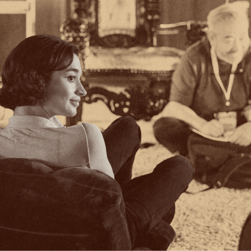 A woman sits in a chair facing a group of people seated on the floor in a casual indoor setting, with a vintage or sepia filter adding an air of longevity to the scene.