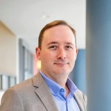 A man in a light gray suit jacket and blue shirt stands indoors near a blue pillar, looking at the camera with a neutral expression.