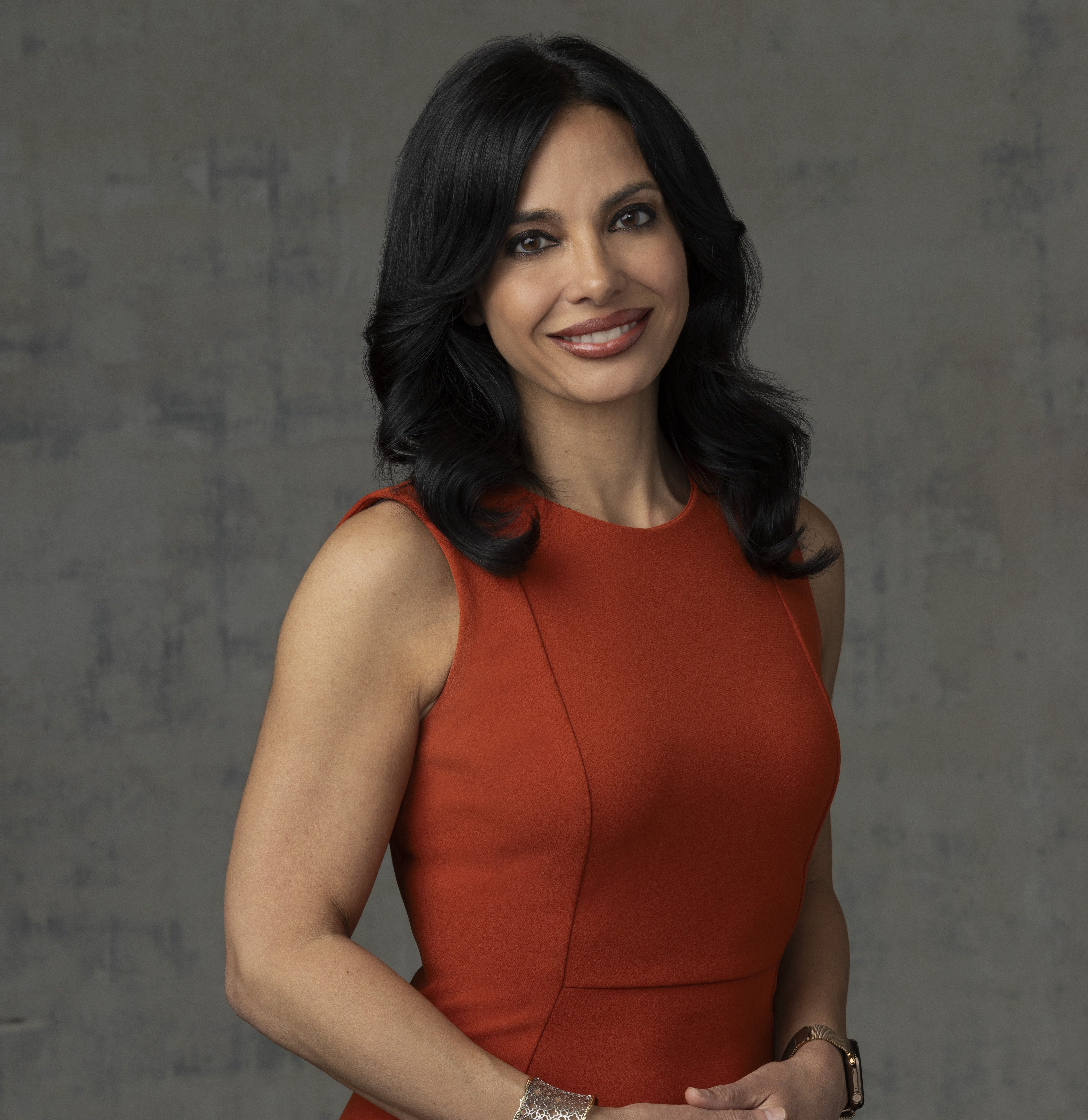 Woman with long dark hair wearing a sleeveless orange dress, silver bracelet, and watch, standing against a gray textured background, smiling at the camera.